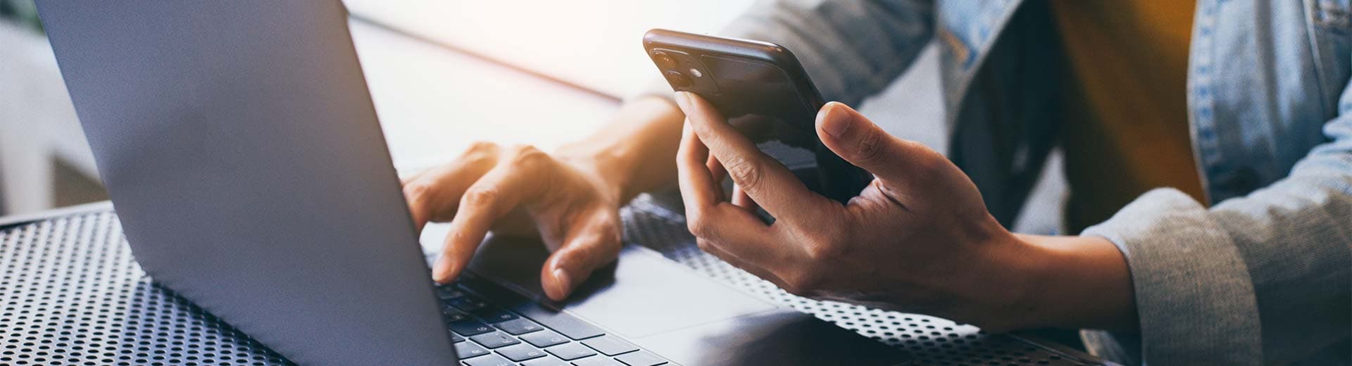 Eine Person sitzt an einem Schreibtisch und bedient einen Laptop und hat ein Mobiltelefon in der Hand.