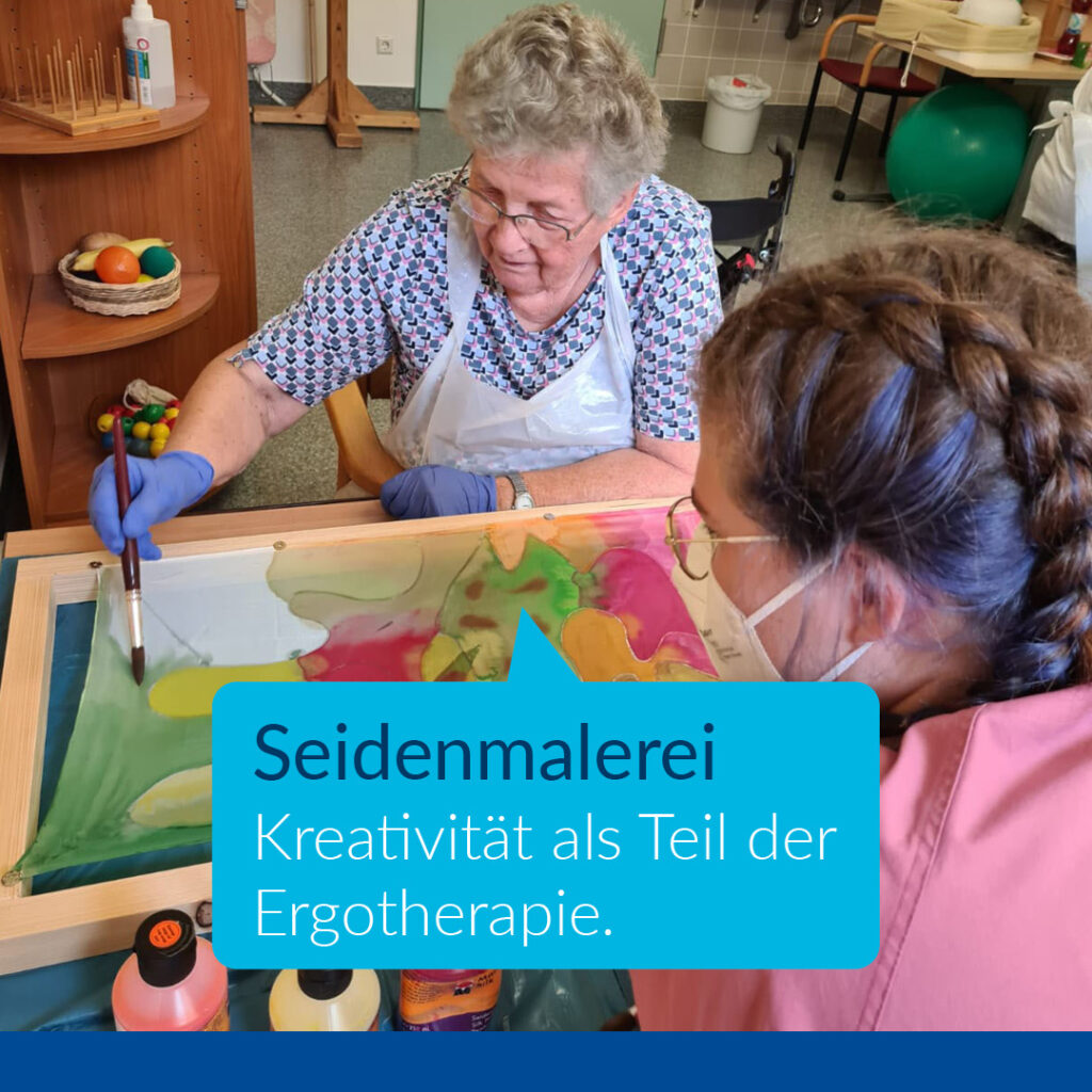 Auf dem Bild ist eine ältere Patientin und eine Pflegerin zu sehen. Die Patientin malt mit bunten Farben auf Seide. Darunter befindet sich ein Text: " Seidenmalerei. Kreativität als Teil der Ergotherapie."